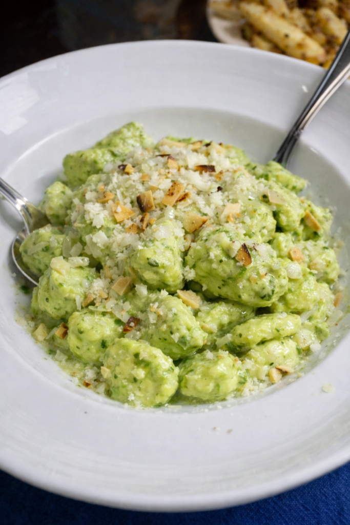 Close-up macro shot of pillowy gnocchi coated in creamy ricotta-basil pesto, wilted baby spinach ribbons and fine lemon zest sprinkled over, micrograted Parmesan snow, tiny pine nut pieces visible in the sauce; silky texture highlighted, shallow depth of field, warm ceramic bowl, gentle backlight to accent creaminess.